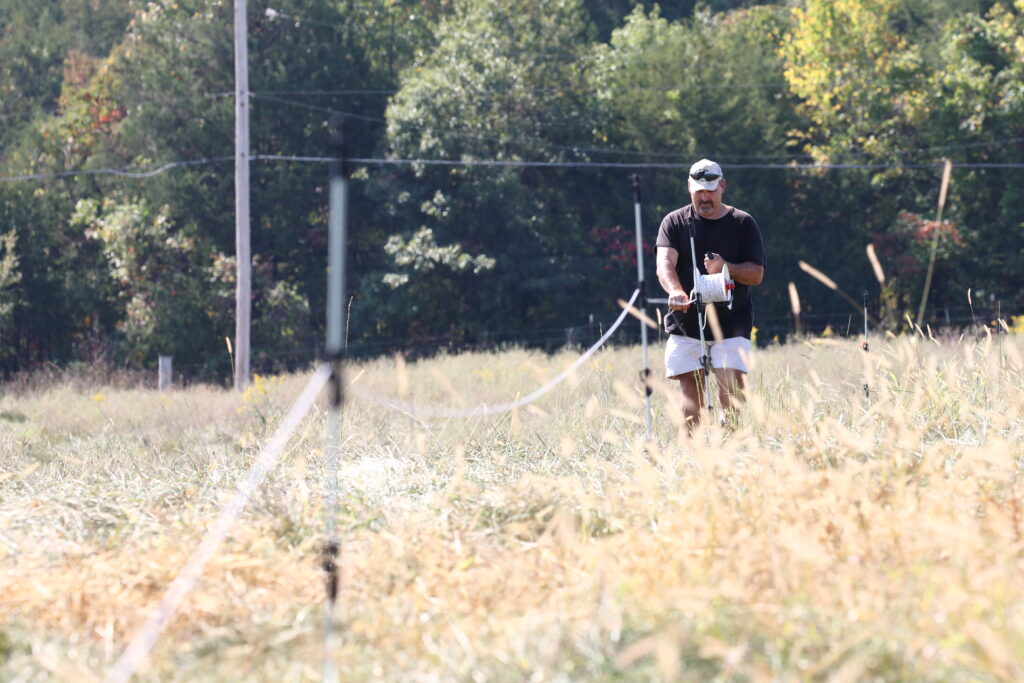 A man reels a spool of poly wire in a field with tall grass in the summertime