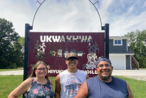 Three individuals are standing in front of a maroon sign that reads "UKWAKHWA" with intricate designs of cornstalks and traditional symbols. The backdrop features a modern farmhouse-style building. The individuals are smiling, conveying a welcoming and friendly atmosphere.