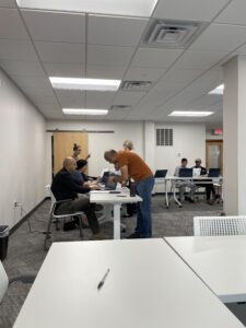 Students in a classroom are seated at tables with laptops, receiving assistance from instructors on basic computer skills.