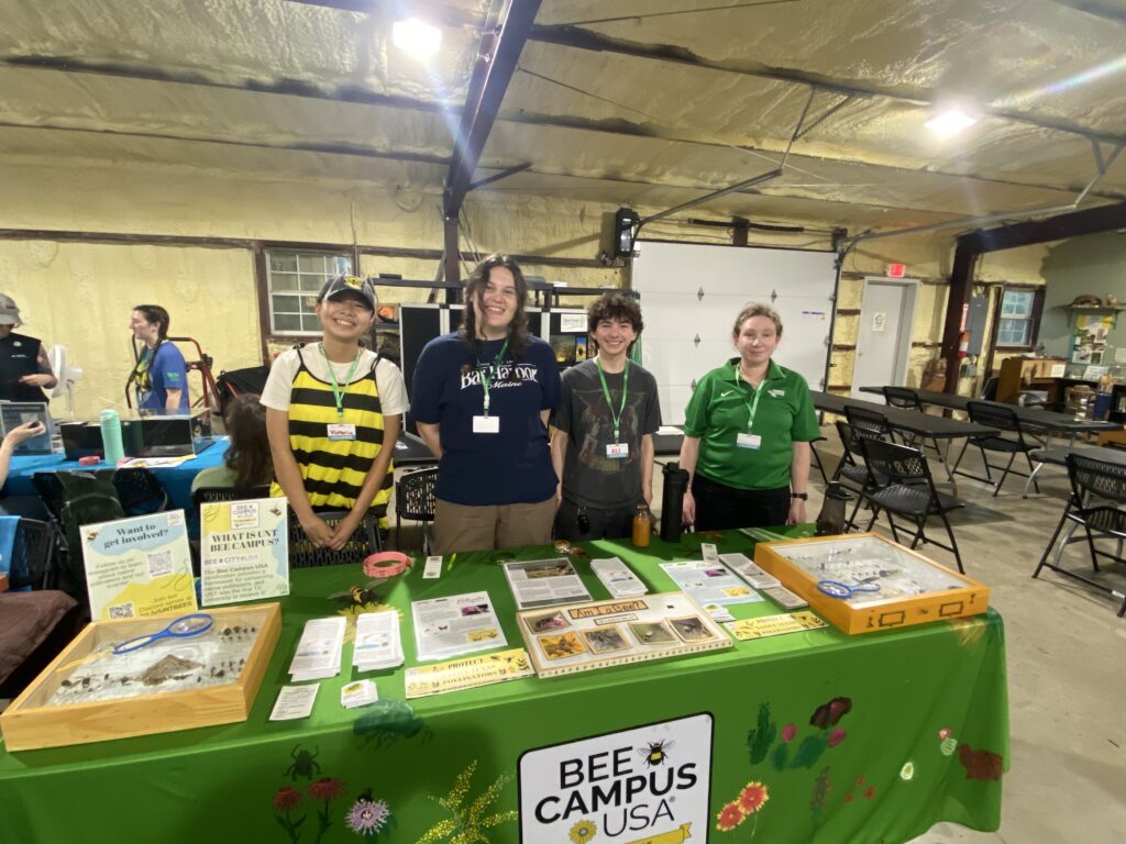 Four people standing behind a table that bears insect display boxes and various informational materials about pollinators