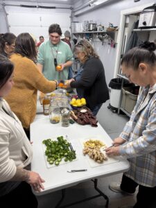 A group of individuals gathered around a table in a well-lit kitchen, actively participating in food preparation. On the table, there are cutting boards with chopped ginger and jalapeños, jars, and various ingredients like oranges and lemons. The participants are engaging in a hands-on workshop.