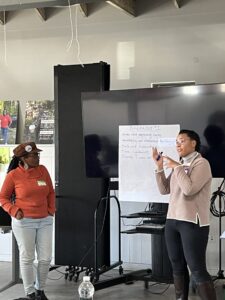 Presenter speaking in front of a sheet that reads: Breakout #1: Nervous about negotiating terms; Accountability and relationship building; Scale and sustainability; time constraints; Diversify Income and… 