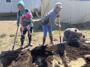 Three women, one with her baby, spread soil and amendments in a raised garden bed. 
