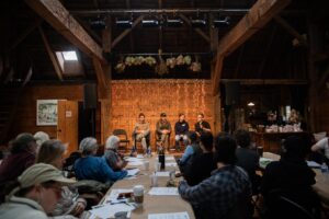 people sitting at tables looking at 4 people on a stage in a panel discussion in an old barn