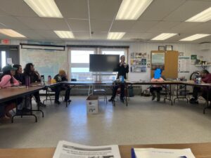 A group of people sit around tables, watching a standing lecturer