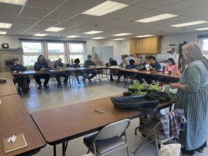 A woman stands in front, showing a couple flats of plant starts to a group of people sitting around a circle of tables. 