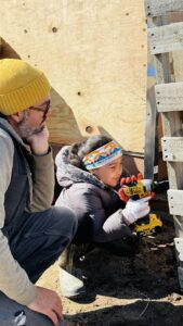 A child drills into the side of an upright pallet structure while an adult watches.