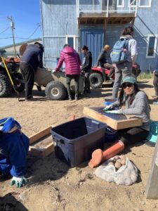 A woman sitting on the ground sifting soil into a plastic tote. There is a bag of potatoes in the foreground and some people standing around a four-wheeler in the background. 