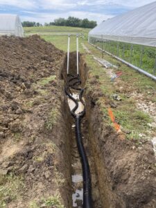 Photo shows a view of a trench alongside a high tunnel. One black corrugated pipe has a wye connection to two smaller pipes, which are attached to PVC pipes sticking up above ground level