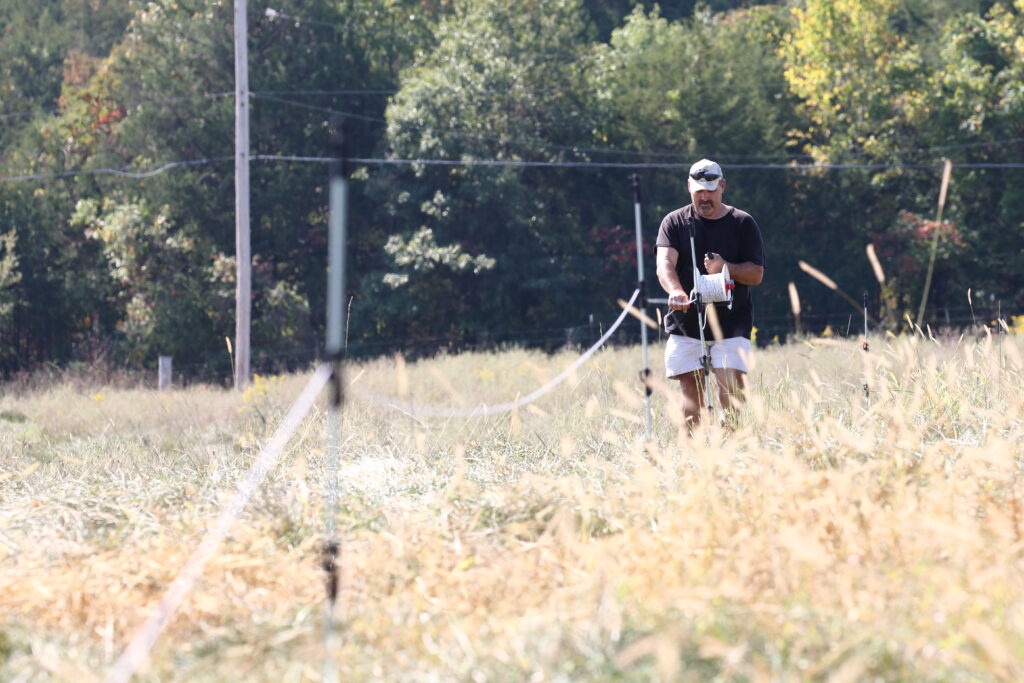 A man reels a spool of poly wire in a field with tall grass in the summertime