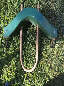 A green bison yoke made of painted wood with a tan bow (bottom loop to hold yoke on neck), lying on grass.