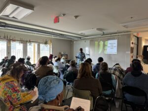 Wil Moss is standing in the front of a packed room next to a powerpoint slide. The lights in the room are off and there is a row of windows on the left. A room full of growers sitting in chairs are looking at Wil. The audience is diverse. 