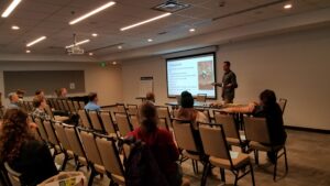 A view of a man presenting in a hotel conference room, taken from the side of the room near the front. Audience members are scattered in rows of chairs, looking away from the camera at his powerpoint slides. 