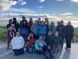 A group of 15 people stand together smiling, with the ocean and sky behind them