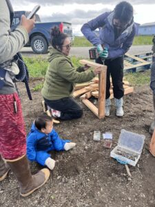 Two young women construct a wooden frame while a baby looks on to the activity