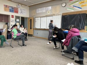 A teacher writes on a white board while several adult learners take notes. 