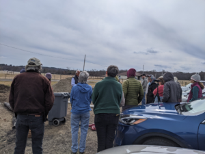 A group of people stand outside on a cloudy winter day at a farm. The group is observing a demonstration on how to build an aerated compost system.